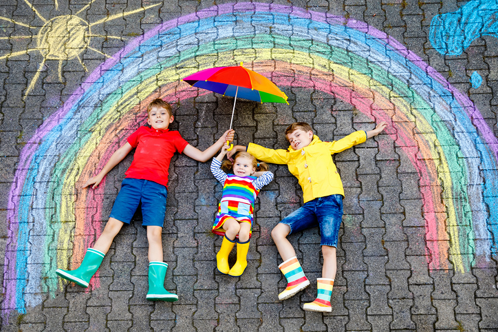 three little children, two school kids boys and toddler girl having fun with with rainbow picture drawing with colorful chalks on asphalt. Siblings in rubber boots painting on ground playing together