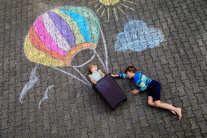 Happy little toddler girl and kid boy flying in hot air balloon painted with colorful chalks in rainbow colors on ground or asphalt in summer. Two children, siblings having fun. Creative leisure.